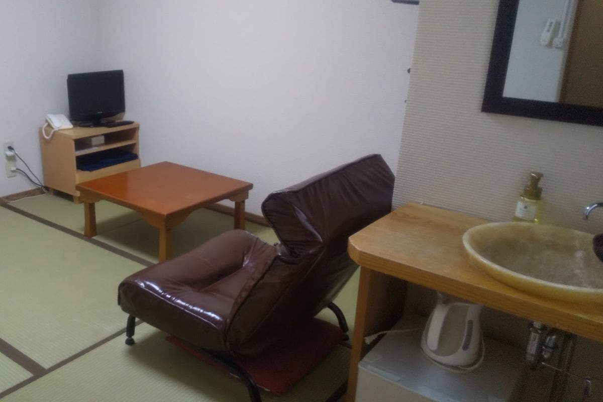 A room with tatami flooring, a wooden table, a leather chair, a television on a stand, and a sink with a mirror.