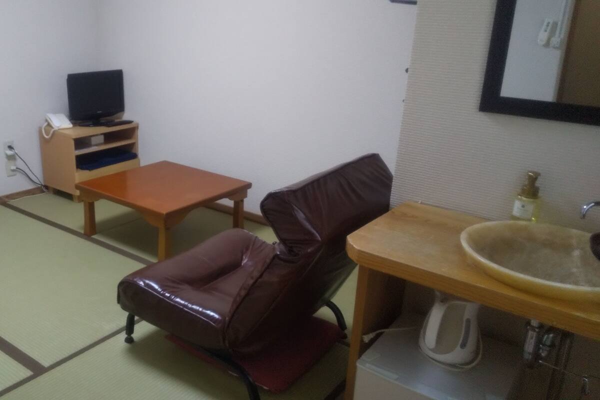 Una habitación con suelo de tatami, una mesa de madera, un sillón de cuero, un televisor sobre un mueble bajo y un lavabo con espejo.