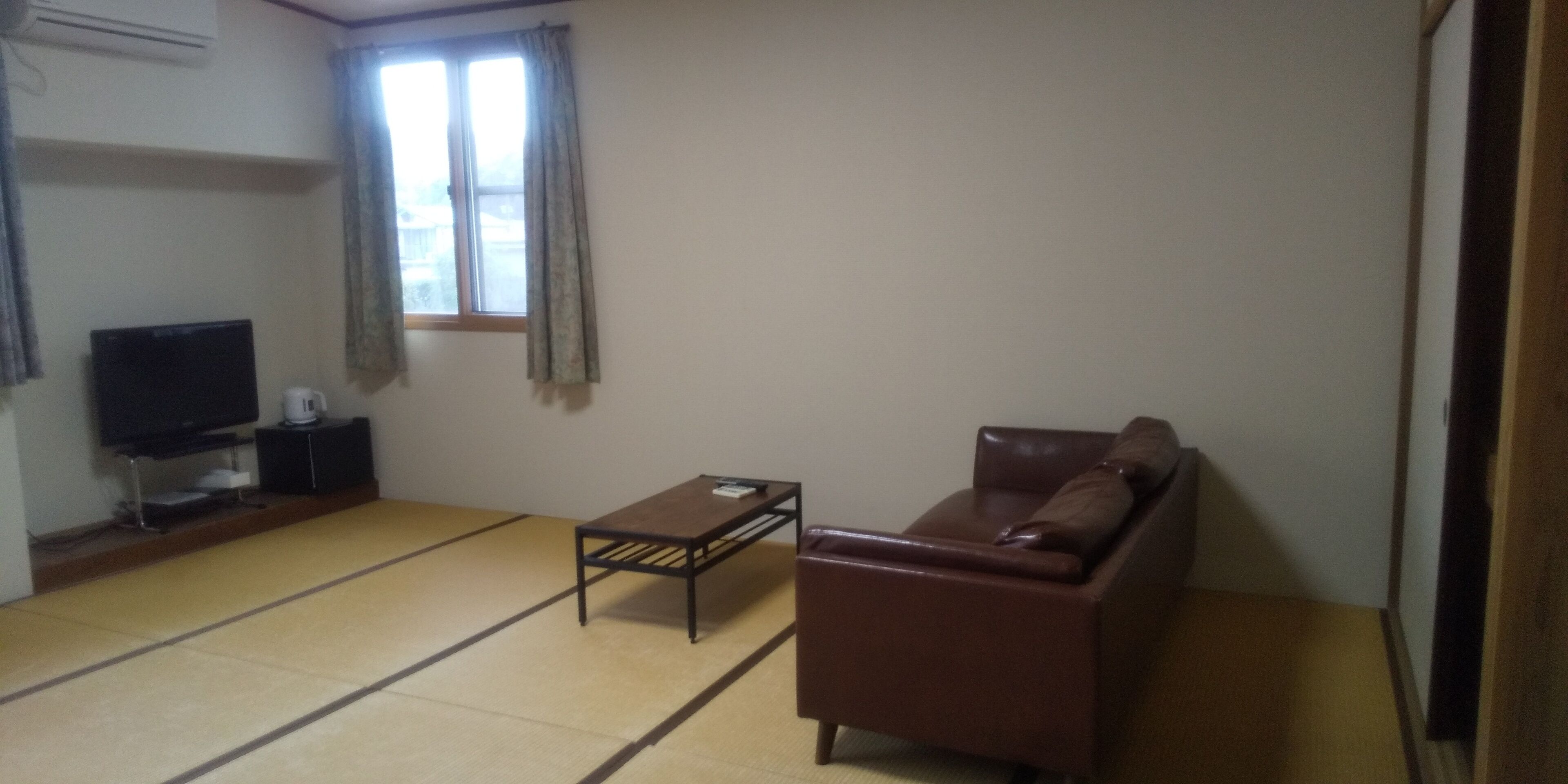 Una sala de estar con suelo de tatami, un sofá de cuero marrón, una mesita, un televisor sobre un mueble bajo y una ventana con cortinas.