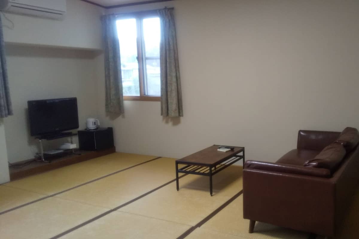 A living room with tatami flooring, a brown leather sofa, a small table, a television on a stand, and a window with curtains.