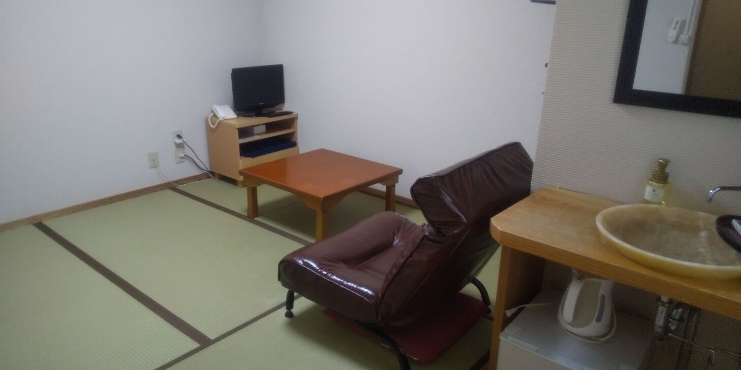 Una habitación con suelo de tatami, un televisor sobre un soporte de madera, un sillón de cuero, una pequeña mesa de madera y un lavabo.