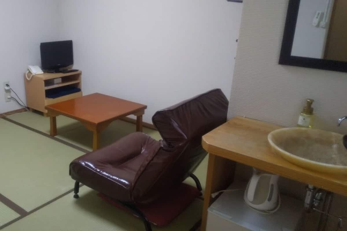A room with tatami flooring, a television on a wooden stand, a leather chair, a small wooden table, and a sink with a basin.