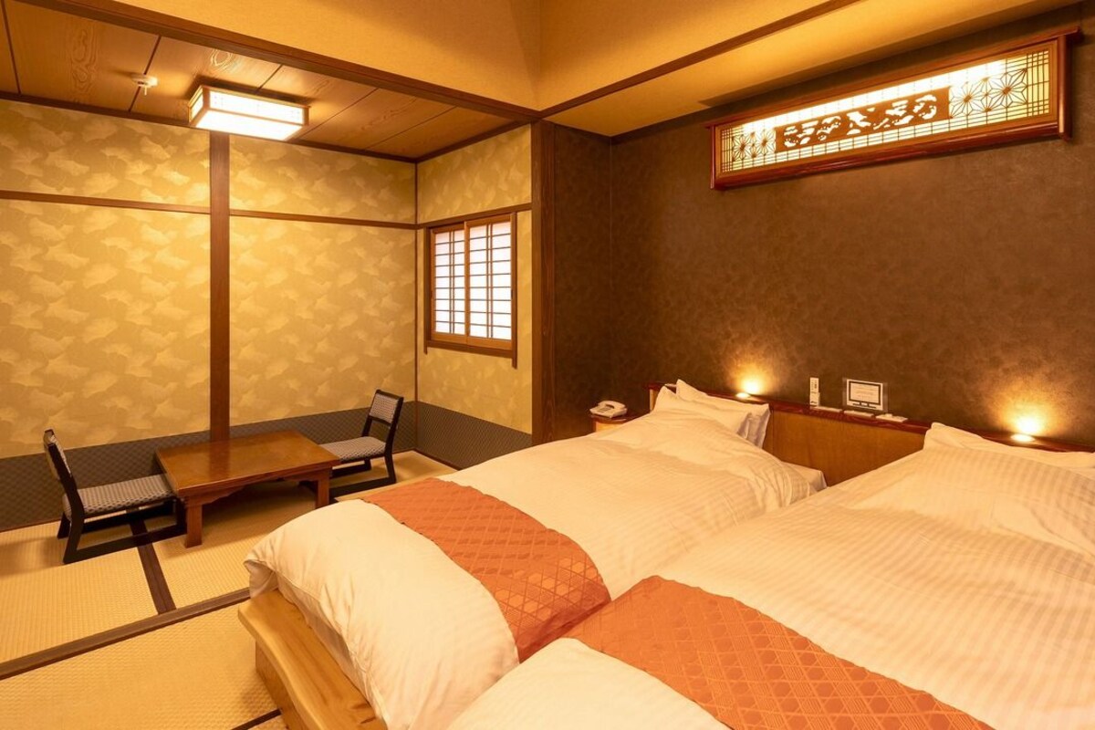 A traditional Japanese-style room with tatami flooring, a low wooden bed with white bedding, a small table, and a chair.