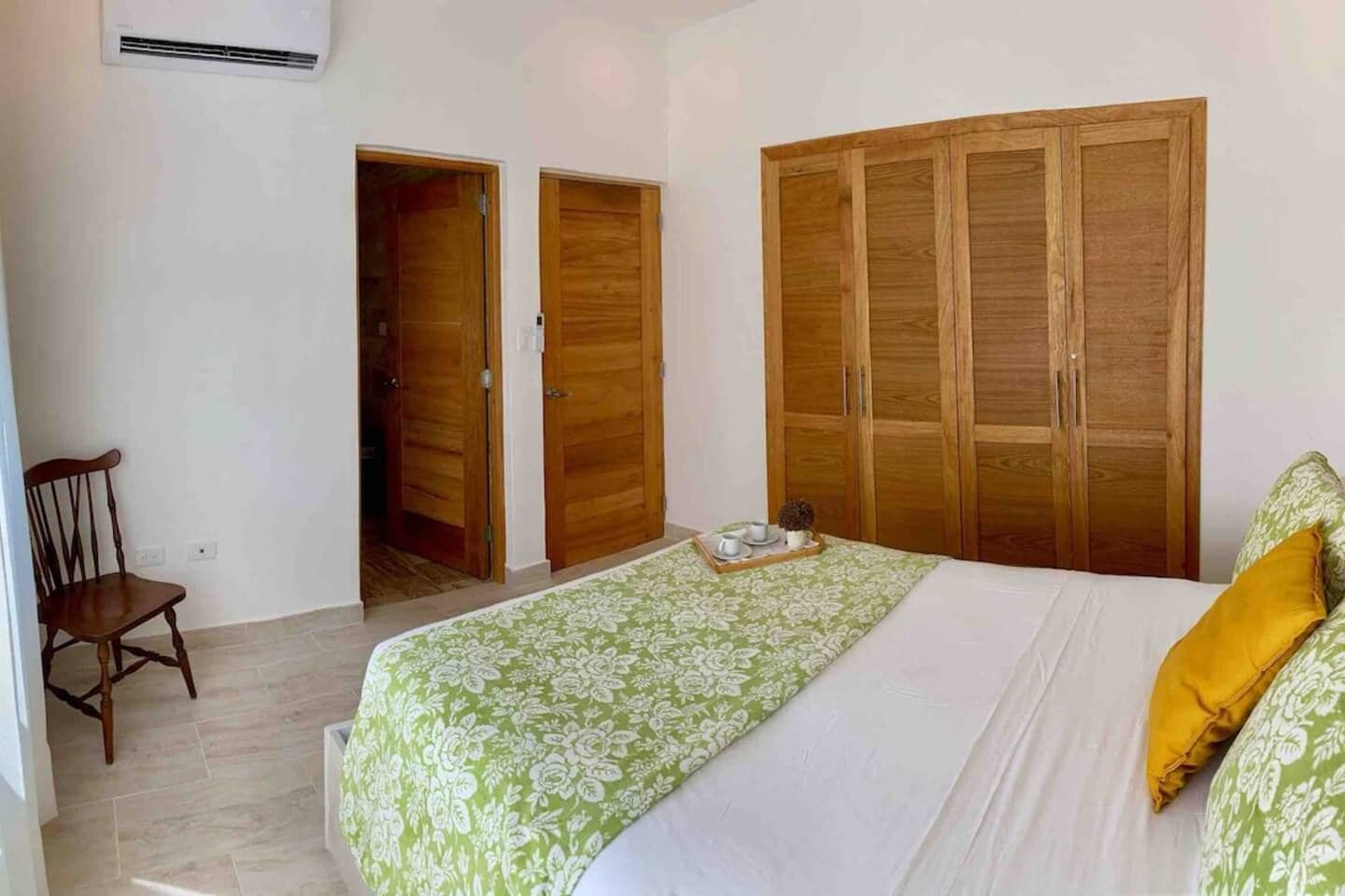 A bedroom with a bed, a wooden wardrobe, a chair, and a small table with a tray.