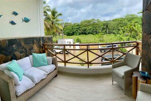A balcony with a stone wall, a wicker sofa with cushions, a wicker armchair, and a view of a parking area and greenery. - ®{Samán~Experience}@Terrenas +Pool+BBQ+5 Min Beach (Las Terrenas)