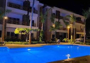 Ein mehrstöckiges Hotel mit Swimmingpool, Palmen und einem geparkten Auto im Hintergrund.