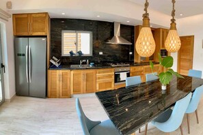 A modern kitchen with a marble countertop, stainless steel appliances, and a dining area with a black table and blue chairs. - ®{Samán~Experience}@Terrenas +Pool+BBQ+5 Min Beach (Las Terrenas)