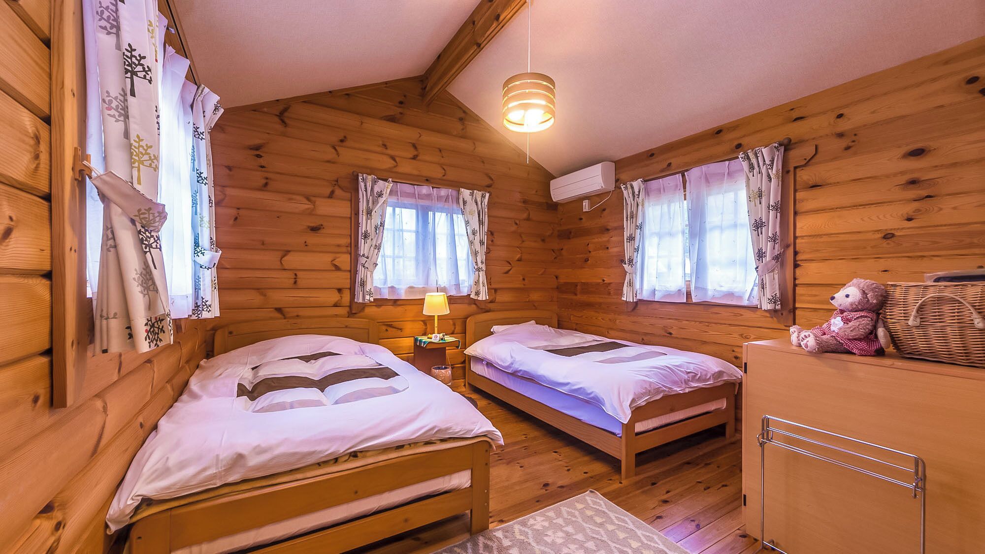 A wooden cabin with two single beds, a nightstand with a lamp, a teddy bear, and a wicker basket.