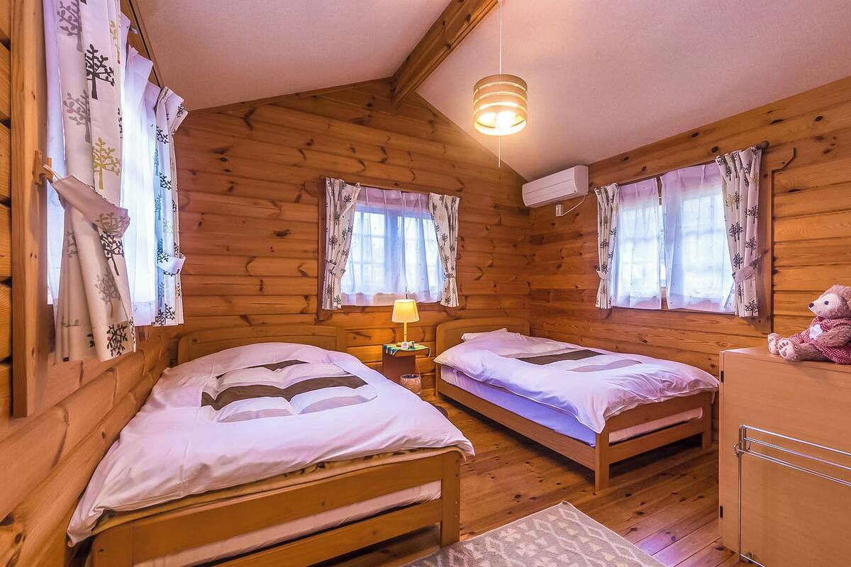 A wooden cabin with two single beds, a nightstand with a lamp, a teddy bear, and a wicker basket.