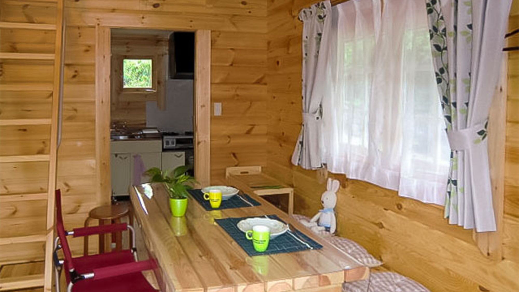 Interior de una cabaña de madera con una mesa de comedor, sillas, una zona de cocina y una ventana con cortinas.