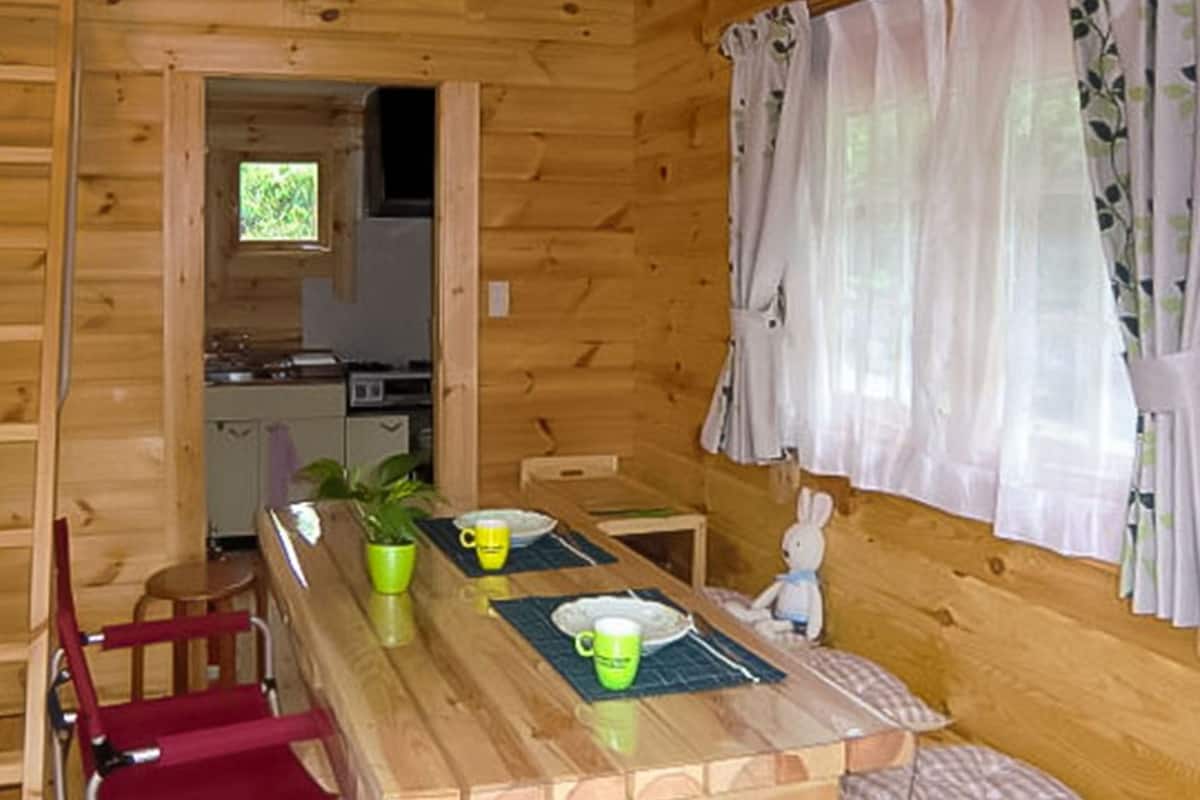 A wooden cabin interior with a dining table, chairs, a kitchen area, and a window with curtains.