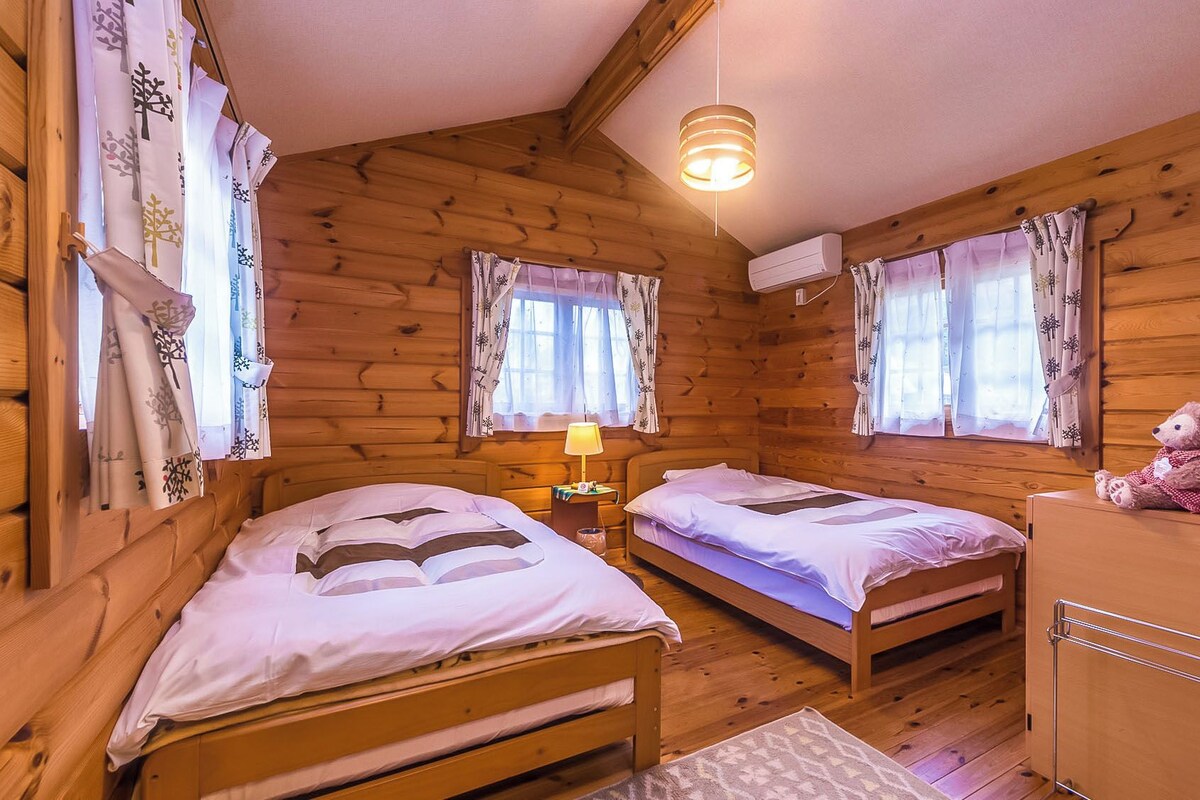 Une cabane en bois avec deux lits simples, une table de chevet avec une lampe, un ours en peluche et un panier en osier.