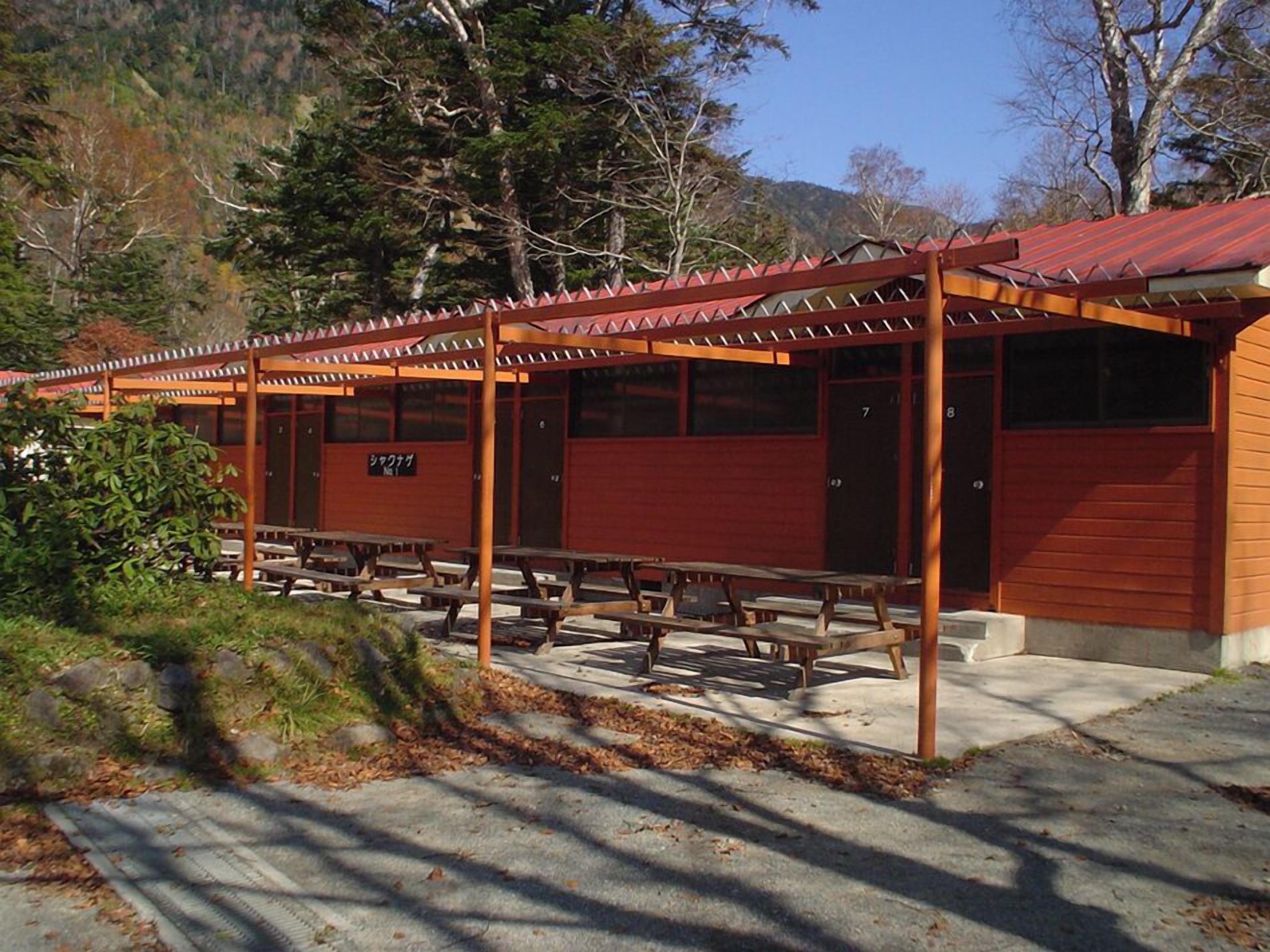 Een overdekt terras met picknicktafels, een houten pergola en een gebouw met een rood dak.