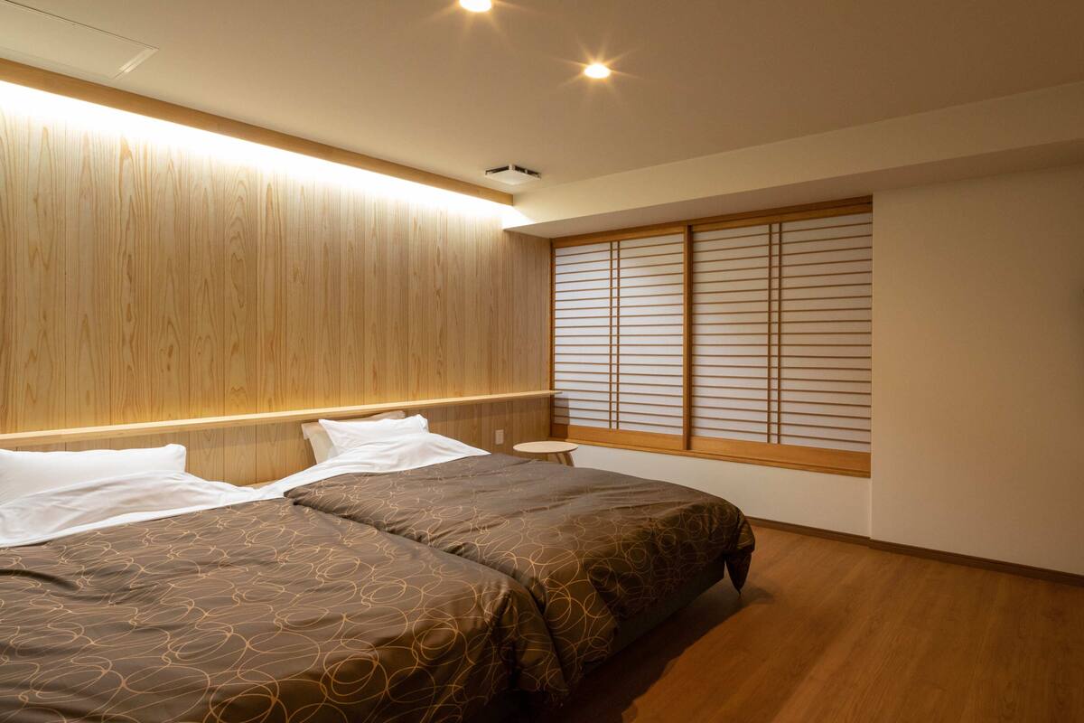 Une chambre à coucher avec un lit, des tables de chevet, une fenêtre avec des stores et une tête de lit en bois.
