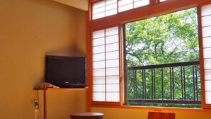 Une pièce de style japonais traditionnel, avec un sol recouvert de tatamis, une table basse, un téléviseur posé sur un meuble et une grande fenêtre donnant sur un paysage verdoyant.