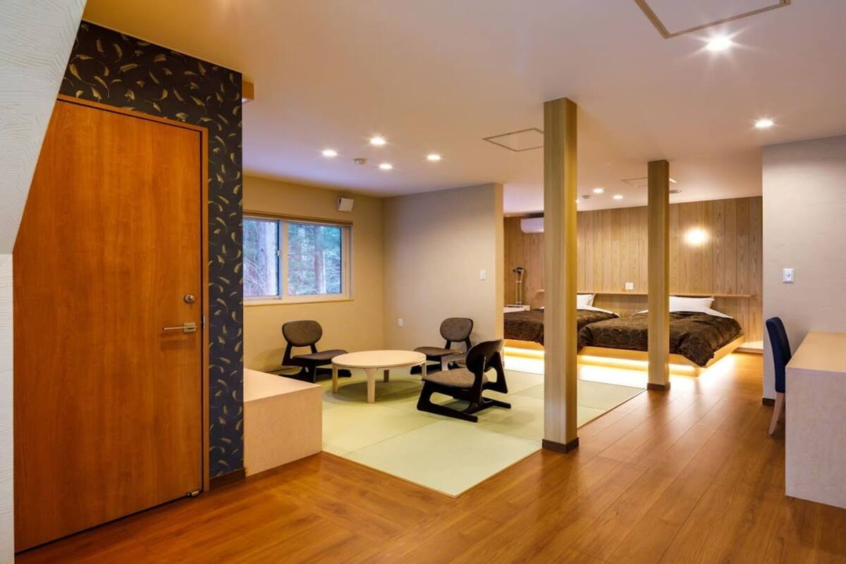 Une chambre d’hôtel moderne avec un plancher en bois, une table à manger ronde, deux chaises, un lit avec une tête de lit en bois, un bureau avec une lampe et une fenêtre donnant sur des arbres.