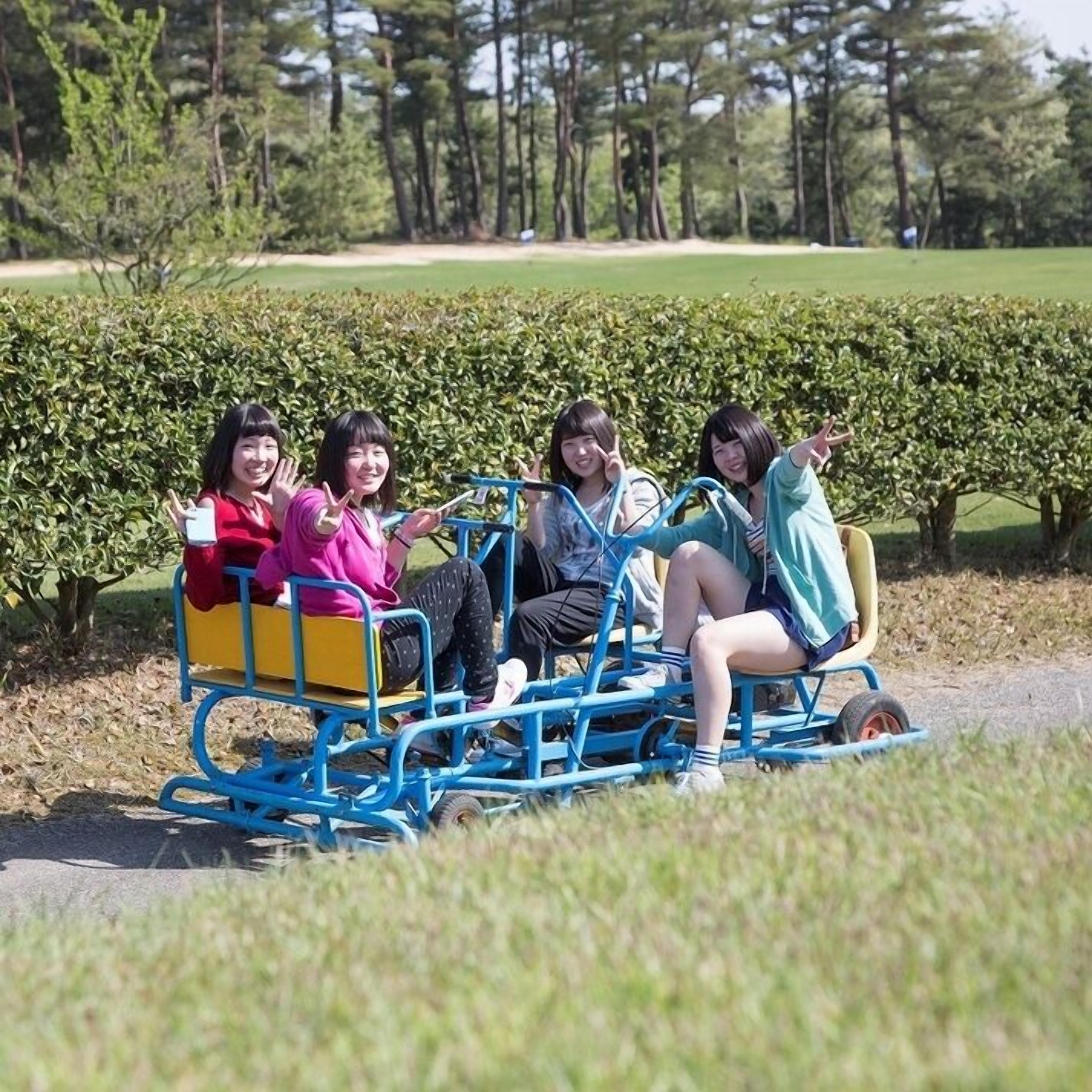 A group of people on a blue cart in a park with trees and a hedge in the background.
