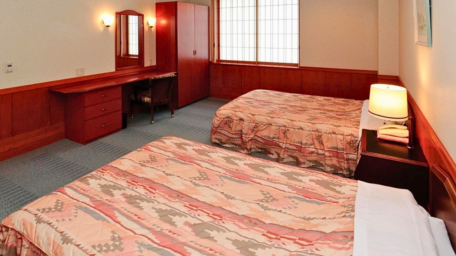 A hotel room with two beds, a desk, a chair, a wardrobe, and a window with blinds.