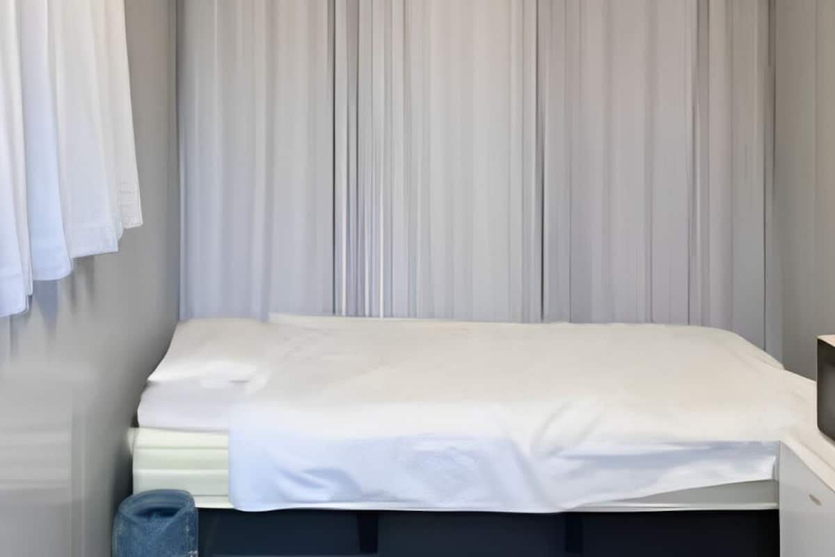 A single bed with a white mattress and sheets, a small bedside table, a wall-mounted TV, and a window with white curtains.