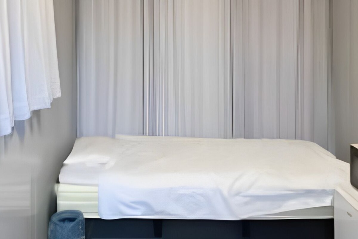 A single bed with a white mattress and sheets, a small bedside table, a wall-mounted TV, and a window with white curtains.