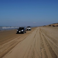 Uma praia com dois carros estacionados na areia, céu claro e ondas do oceano ao fundo.