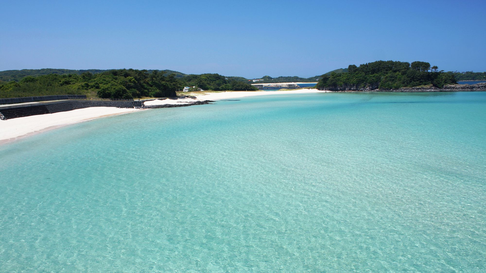 A coastal beach with clear turquoise waters, a sandy shore, and a forested hill in the background.