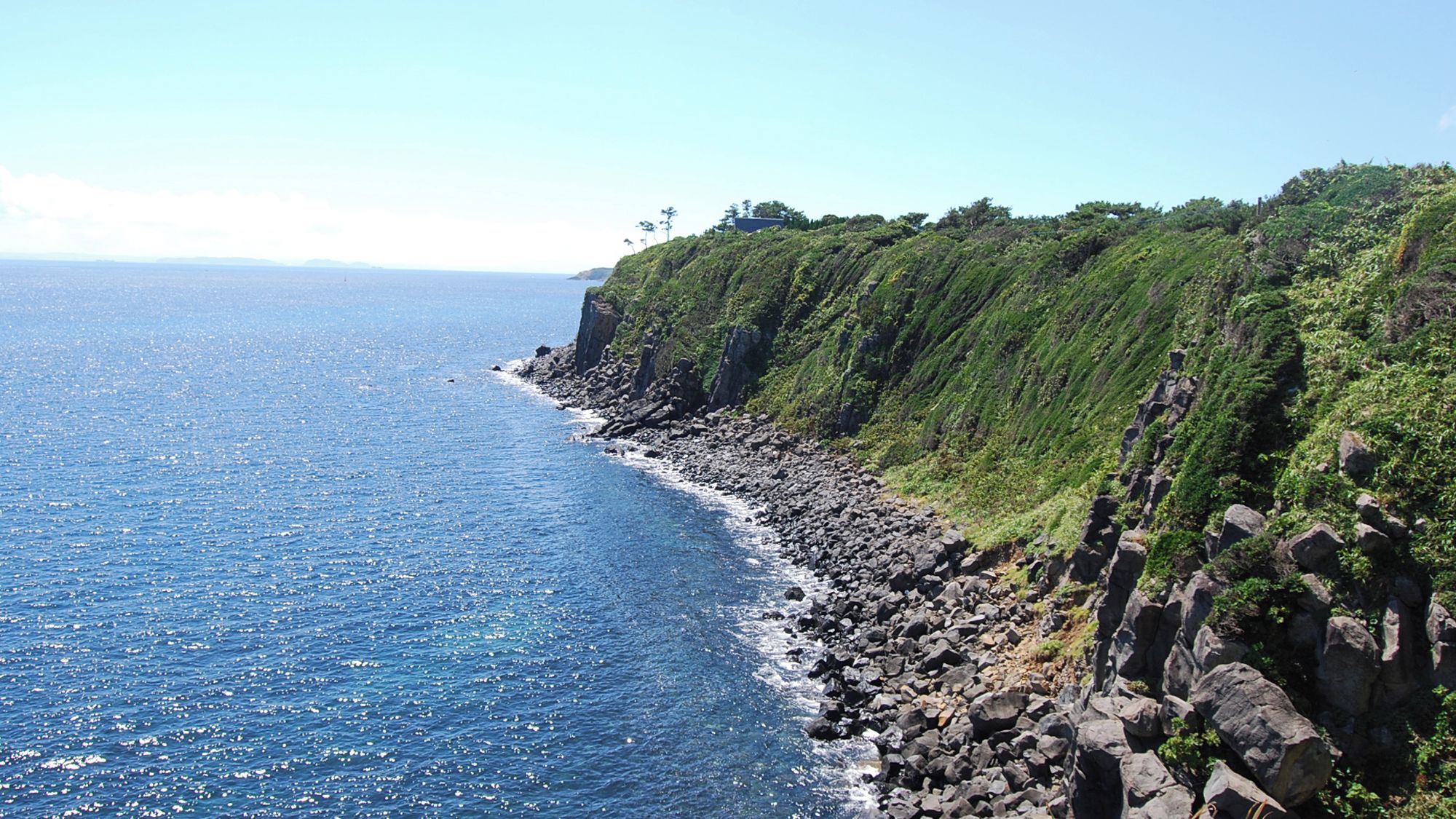 A coastal landscape with a rocky shoreline, a clear blue sea, and a steep cliff with greenery.