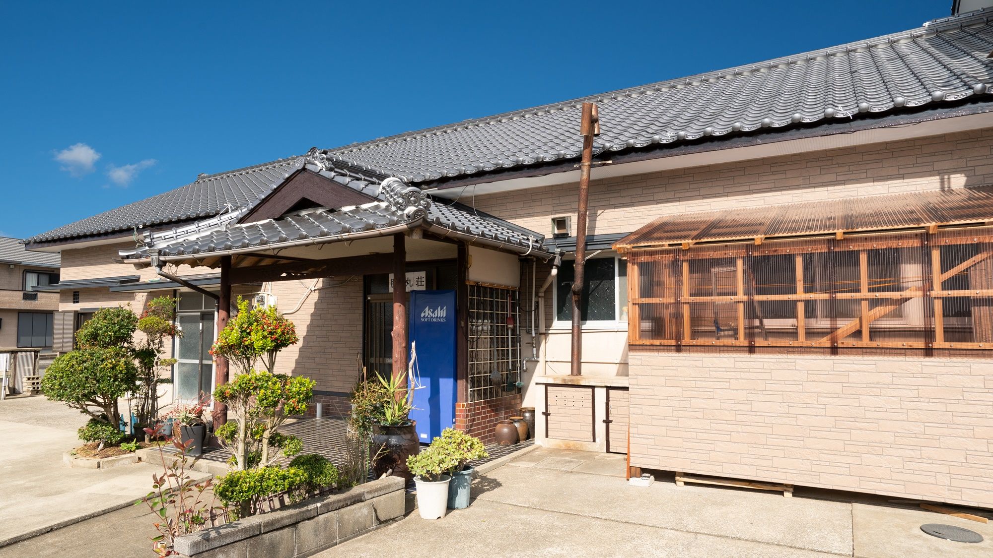 Ein traditionelles japanisches Haus mit einem Ziegeldach, einer Holzterrasse und einem kleinen Garten mit Topfpflanzen.