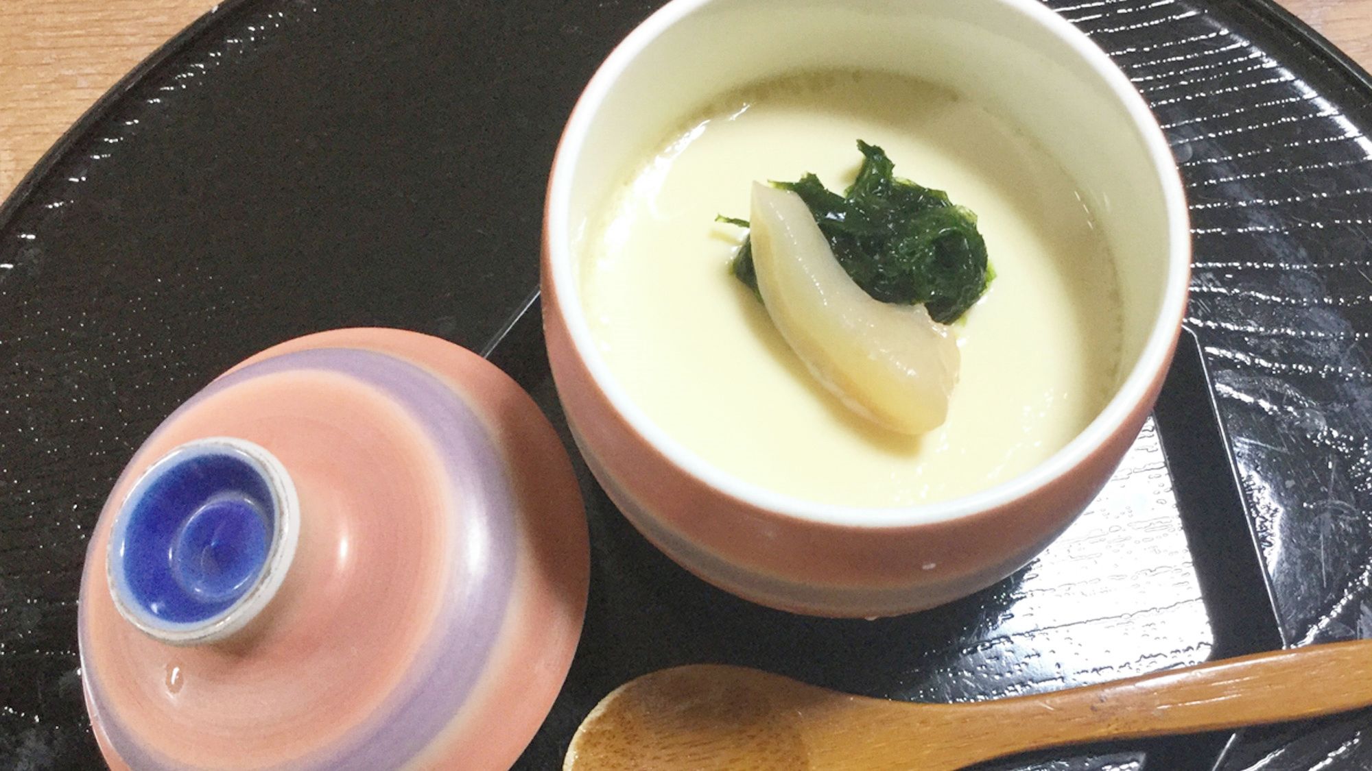 A bowl of soup with a spoon and a bottle on a black tray.