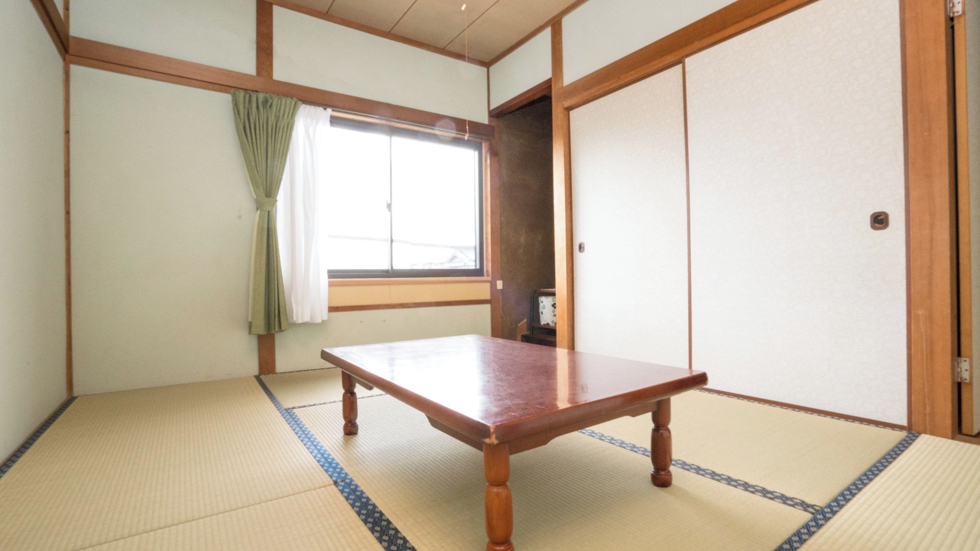 A traditional Japanese room with tatami flooring, a wooden table, a sliding door, and a window with curtains.