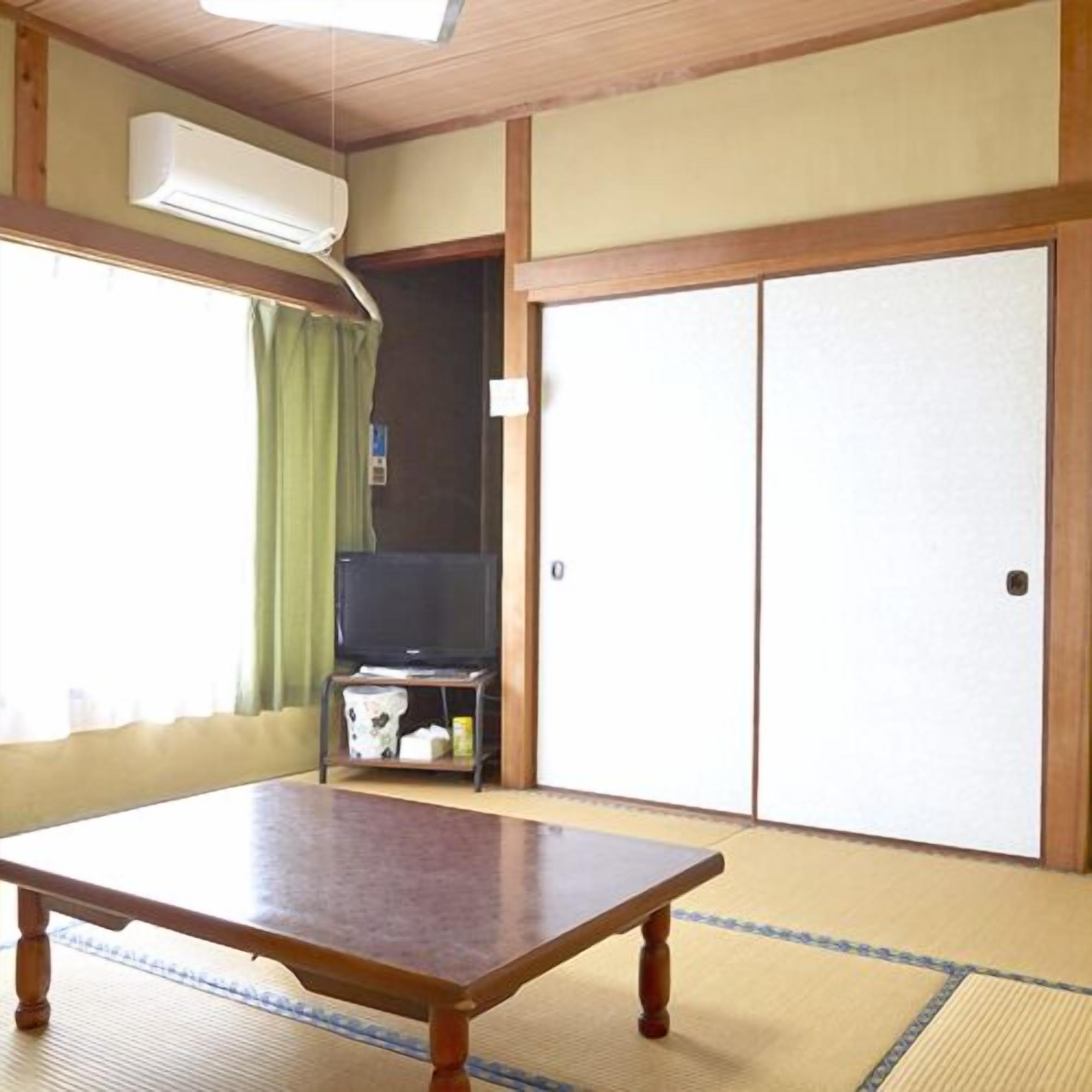 Une pièce avec un sol recouvert de tatamis, une table en bois, un téléviseur posé sur un meuble et des portes coulissantes.