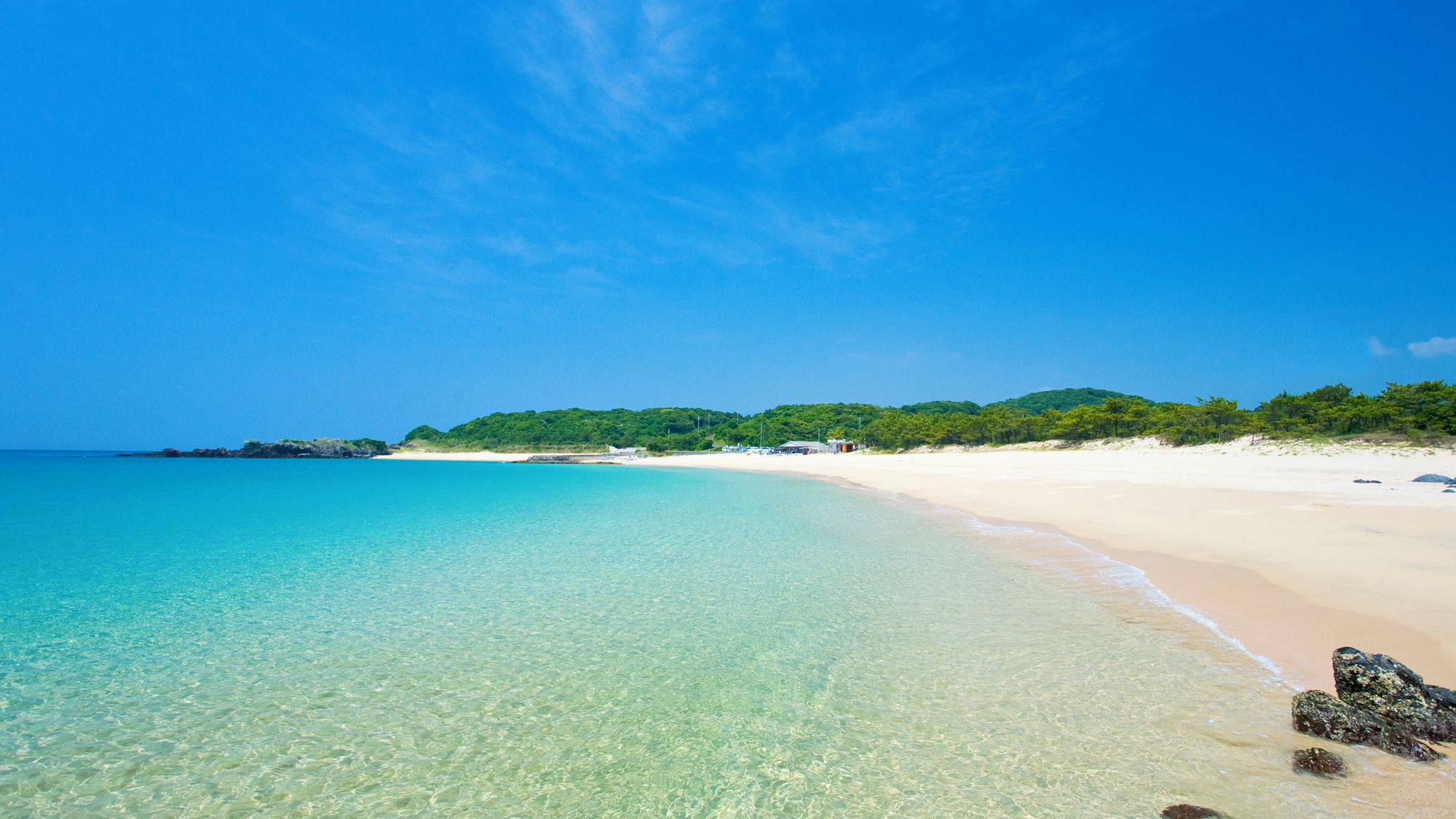 A beach with clear turquoise water, sandy shores, and green hills in the background.
