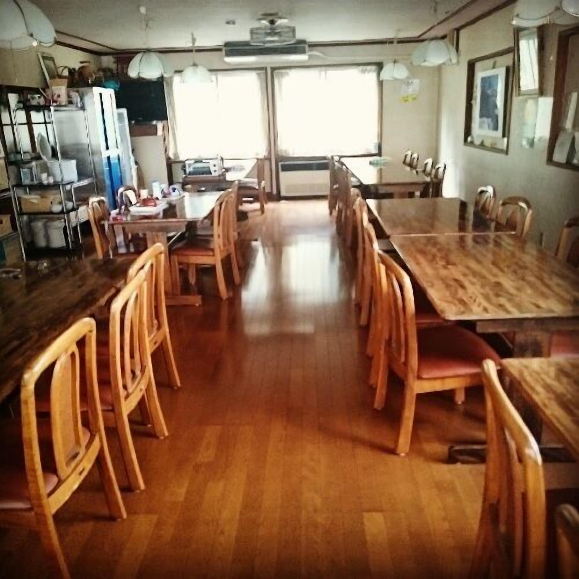 Un comedor con mesas y sillas de madera, una cocina al fondo y ventanas amplias.