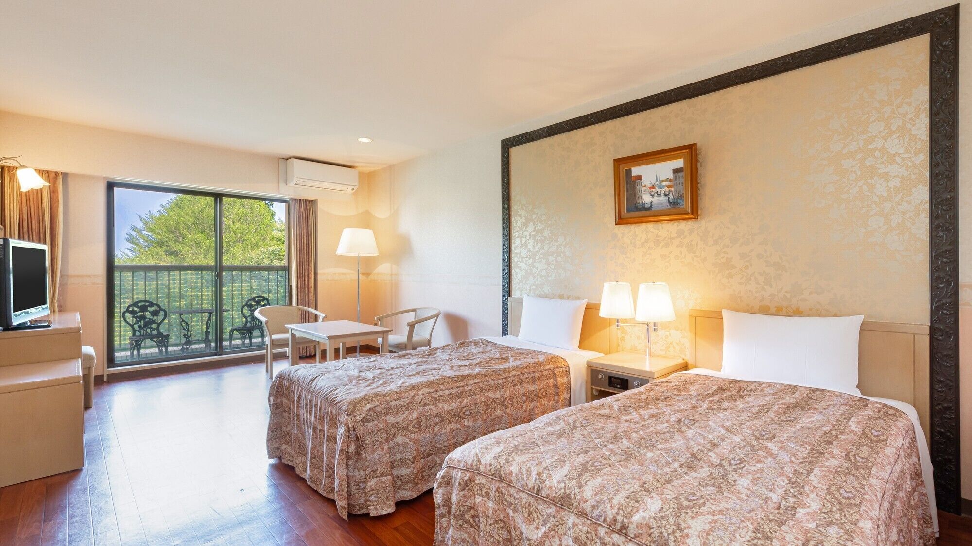 A hotel room with two beds, a desk, chairs, a balcony with a view, and a painting on the wall.