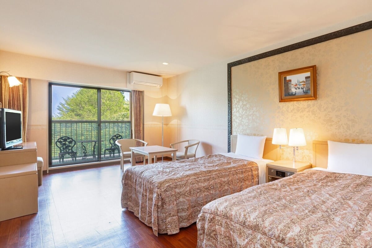 A hotel room with two beds, a desk, chairs, a balcony with a view, and a painting on the wall.