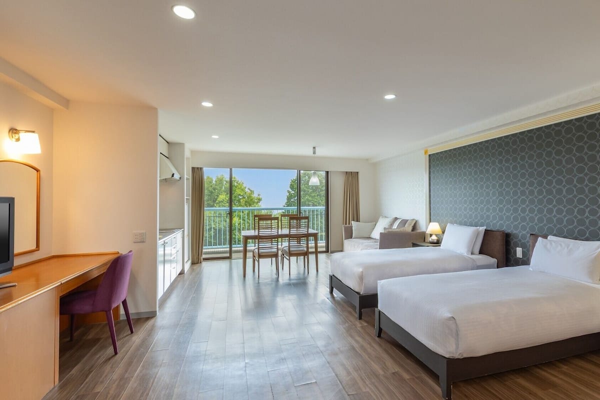 A hotel room with two beds, a desk, a chair, a TV, and a balcony with a view.