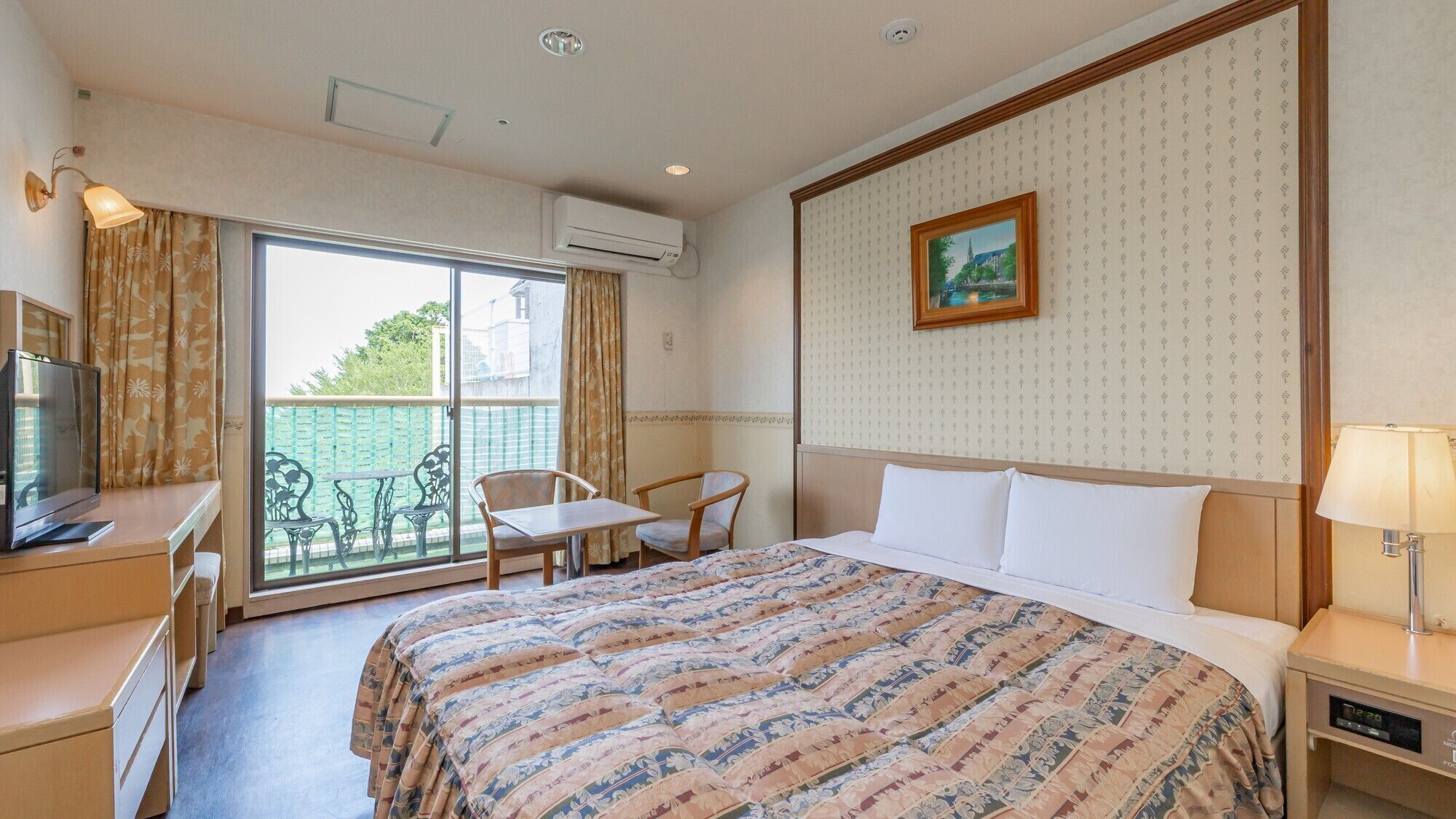 A hotel room with a bed, a desk with a television, a chair, a balcony with a view, and an air conditioning unit.