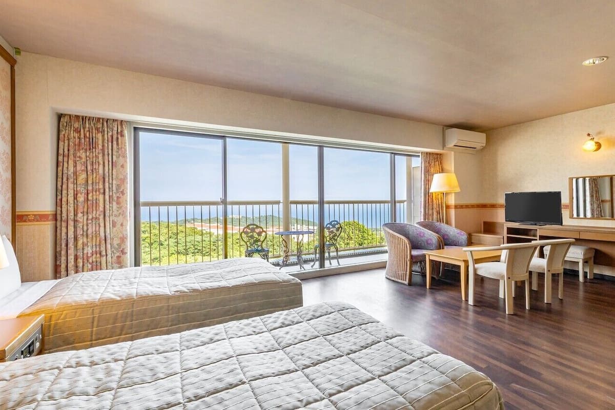 A hotel room with two beds, a desk with a TV, a balcony with a view, and a lamp.