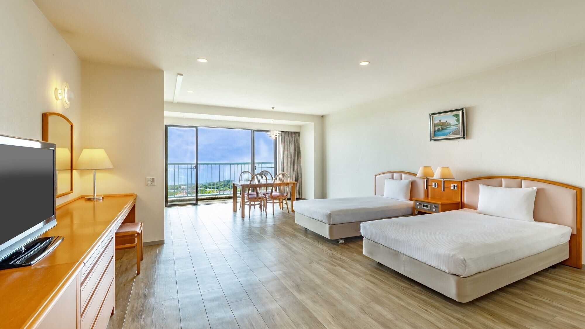 A hotel room with two beds, a desk with a TV, a dining area with chairs, and a large balcony with a view.