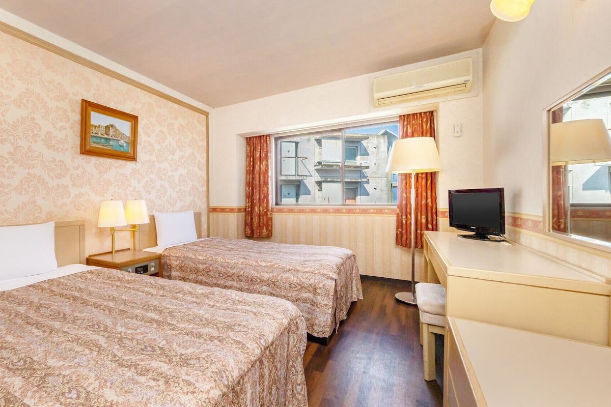 A hotel room with two beds, a desk, a television, and a window with curtains.