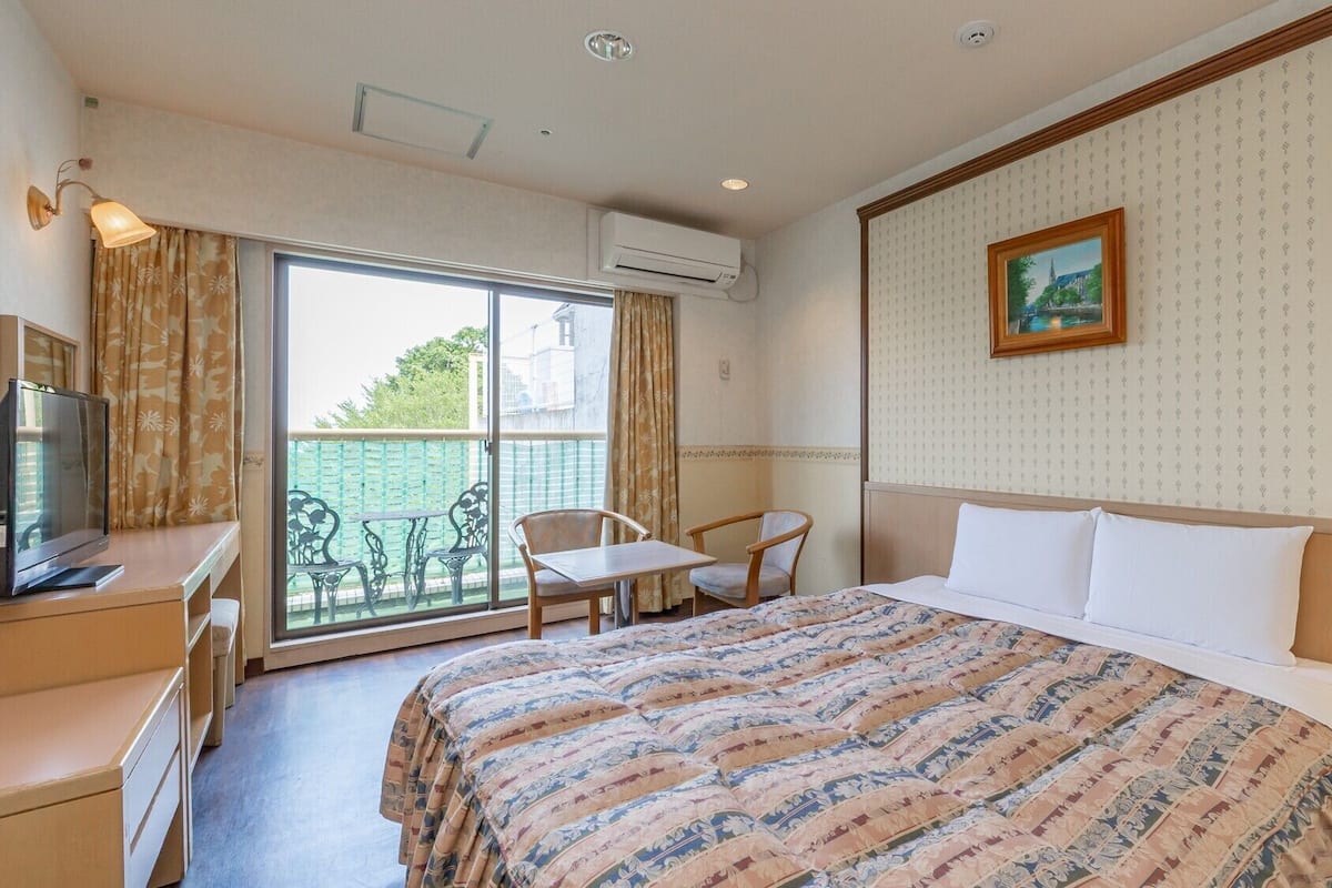 A hotel room with a bed, a desk with a television, a chair, a table, and a balcony with a view.