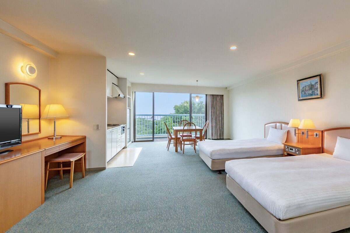 A hotel room with two beds, a desk with a TV, a balcony with a table and chairs, and a painting on the wall.