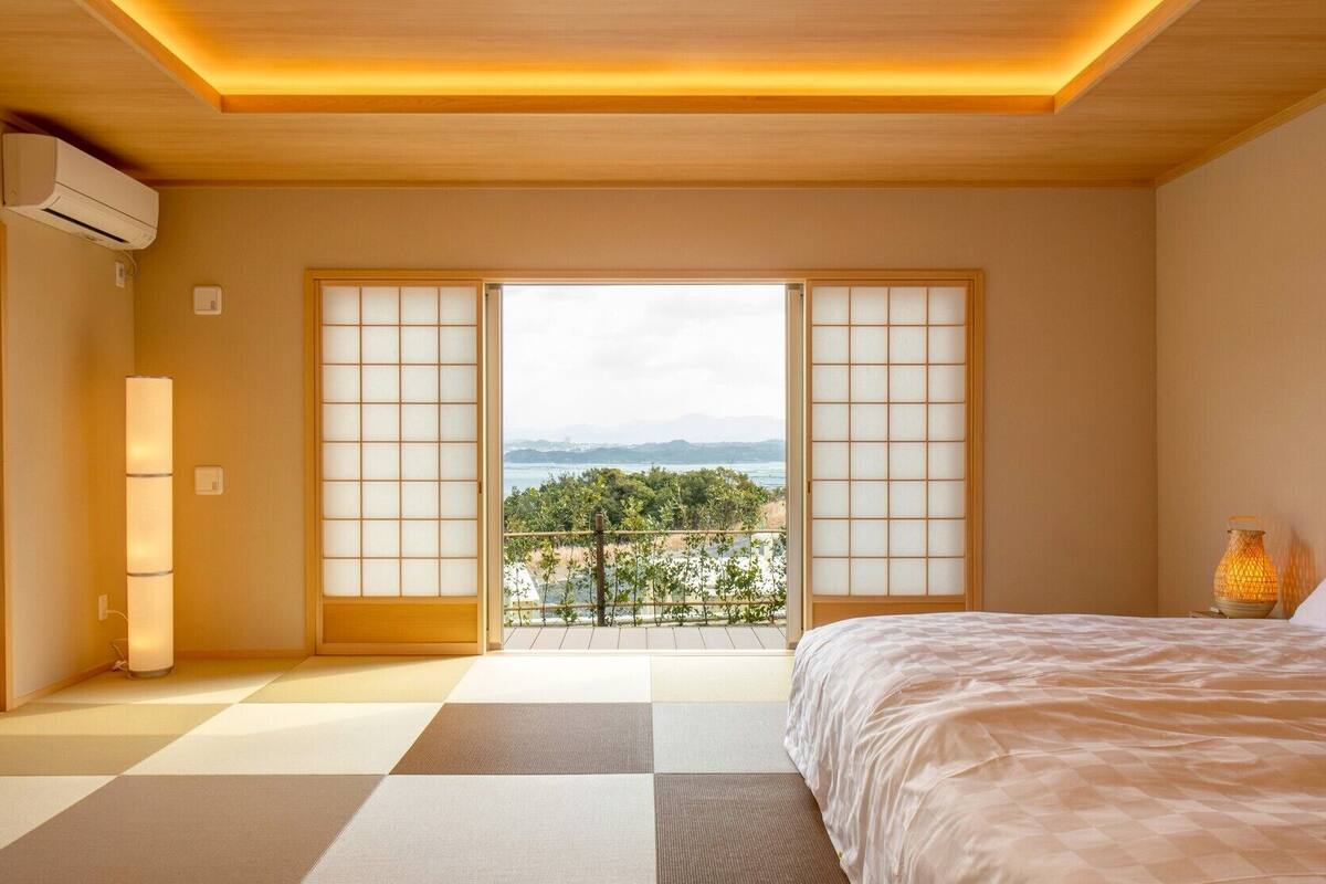 Une chambre d’hôtel avec un sol recouvert de tatamis, un lit, une table de chevet avec une lampe, et une vue sur un jardin et des montagnes.