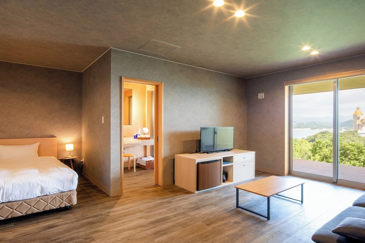Une chambre d’hôtel moderne équipée d’un lit, d’un bureau avec une télévision, d’un petit tabouret et offrant une vue sur un château grâce à de grandes fenêtres.