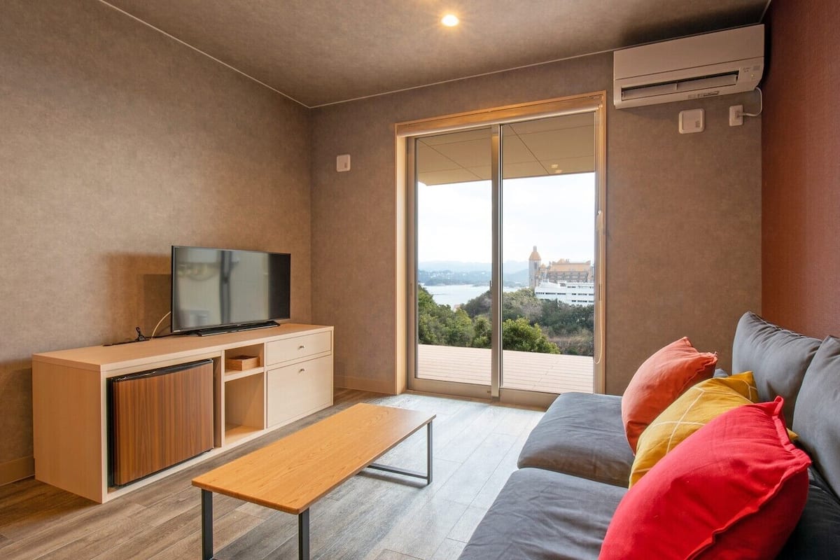 Un salon moderne comprenant un canapé, un téléviseur, un banc et une porte-fenêtre coulissante donnant sur un balcon avec vue sur la ville.