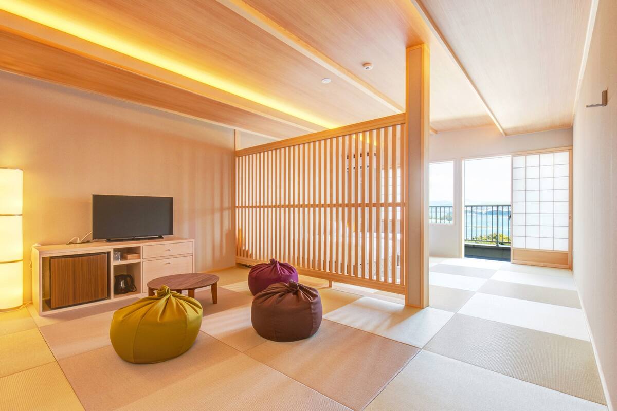 Une chambre d’hôtel moderne dotée d’un sol en tatami, d’une télévision, d’une cloison en bois et offrant une vue sur l’extérieur.