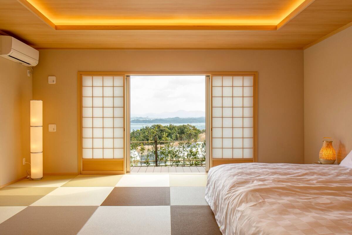 Une pièce dotée d’un sol recouvert de tatamis, d’un lit, d’une table de chevet avec une lampe, et offrant une vue sur la verdure et les montagnes à travers des panneaux coulissants.