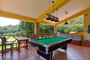 A pool table with a green felt surface, surrounded by wooden chairs and a bench, set on a balcony with a view of a lush green landscape.
