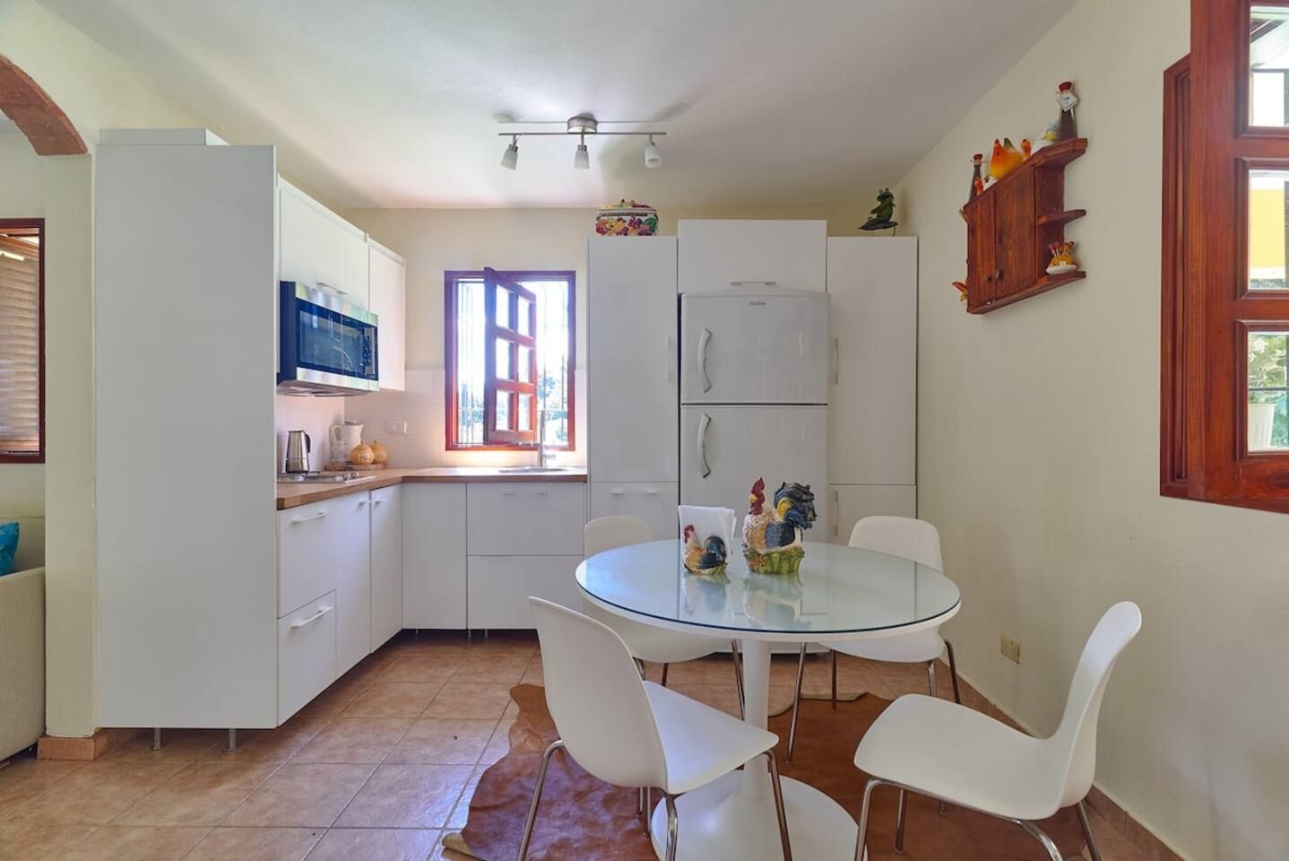 Una cocina compacta con armarios blancos, una mesa redonda de cristal y sillas blancas.