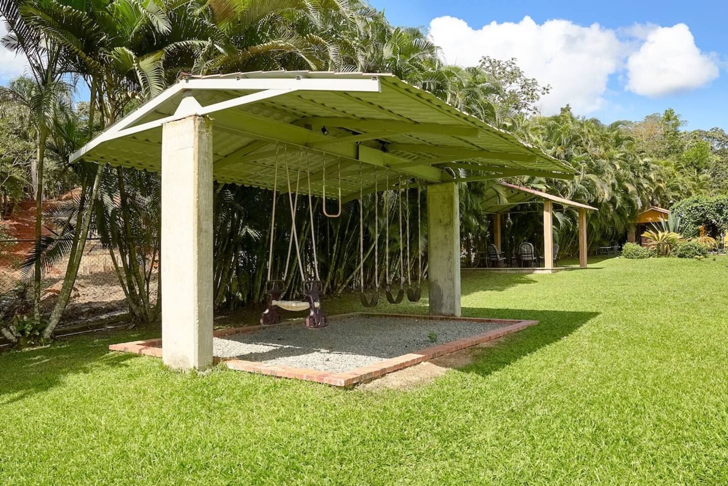 Un espacio de estar al aire libre con techo, con columpios, rodeado de abundante vegetación y palmeras.